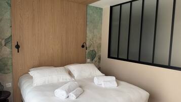 A neatly made bed with white bedding and two rolled towels on top, a wooden wardrobe, and a wall-mounted light fixture.