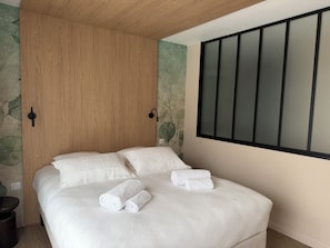 Una cama bien tendida con ropa blanca y dos toallas enrolladas encima, un armario de madera y una lámpara fijada a la pared.