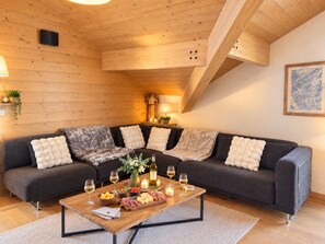A cozy living room with a dark grey sectional sofa, a wooden coffee table with snacks and drinks, and a wooden ceiling with exposed beams. - Le Jardin Alpin A 41 - Les Balcons de Suzanne (Vaujany)