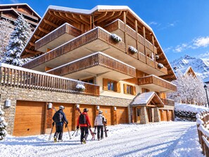 A multi-story wooden building with balconies, ski equipment, and snow-covered surroundings. - Le Jardin Alpin A 41 - Les Balcons de Suzanne (Vaujany)