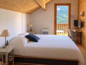 A bedroom with a bed, bedside tables, a desk with a chair, a TV, and a balcony with a view of trees. - Le Jardin Alpin A 41 - Les Balcons de Suzanne (Vaujany)