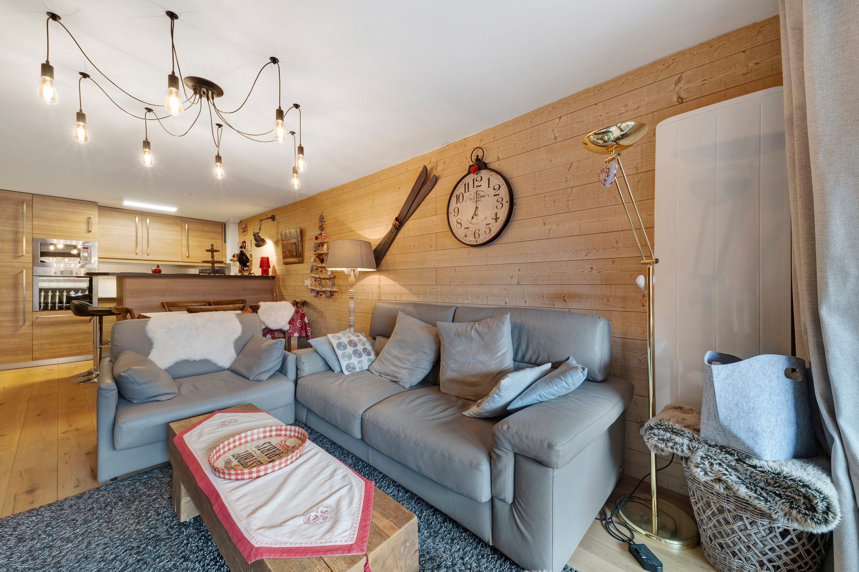 A modern living room with a grey sofa, a wooden wall, and a kitchen in the background.