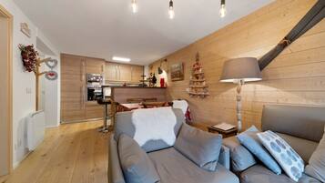 Een moderne woonkamer met een grijze bank, houten vloeren, een keuken op de achtergrond en een kerstboomversiering.