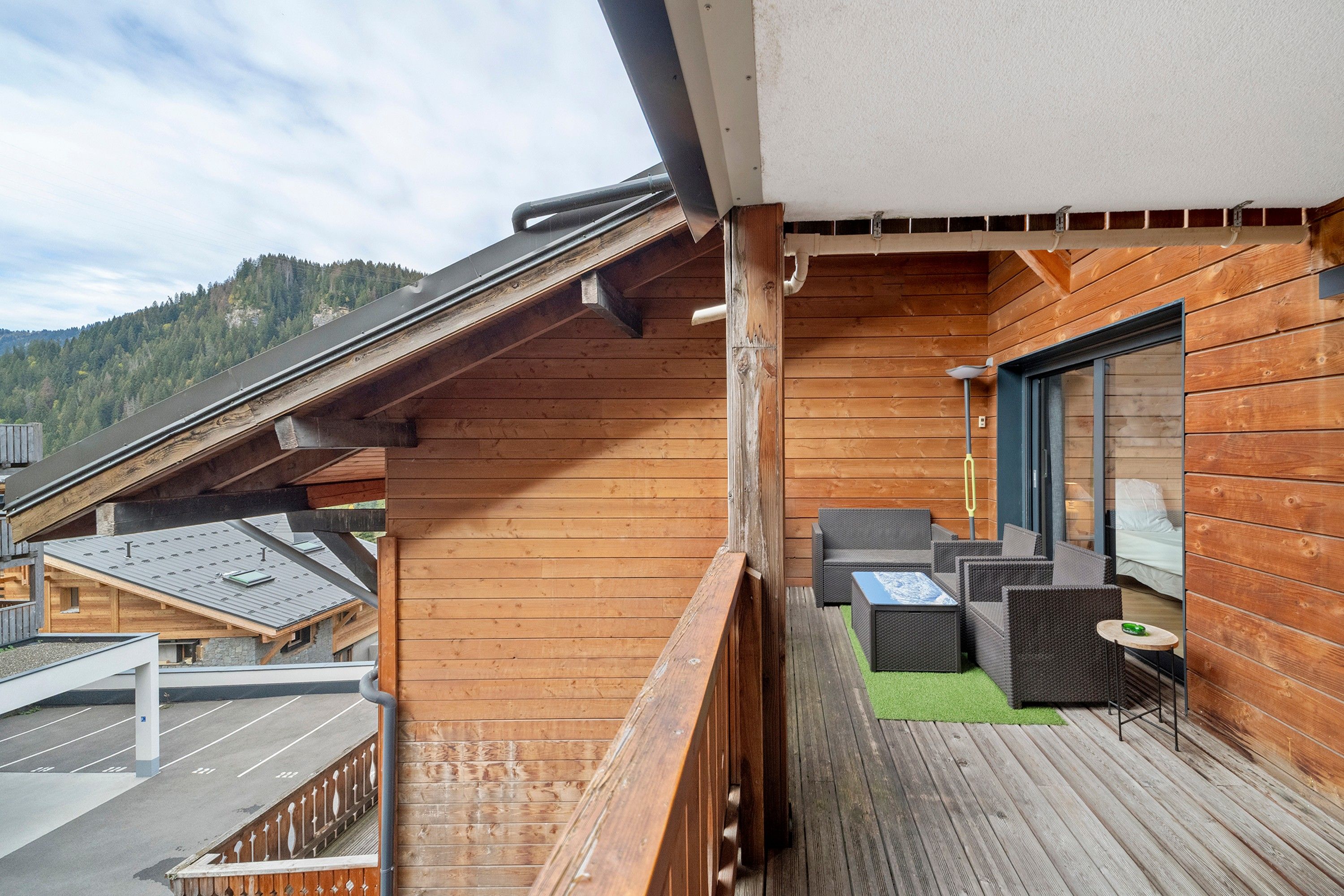 Ein Holzbalkon mit Gartenmöbeln, Blick auf einen bewaldeten Berg und ein Gebäude mit Satteldach.