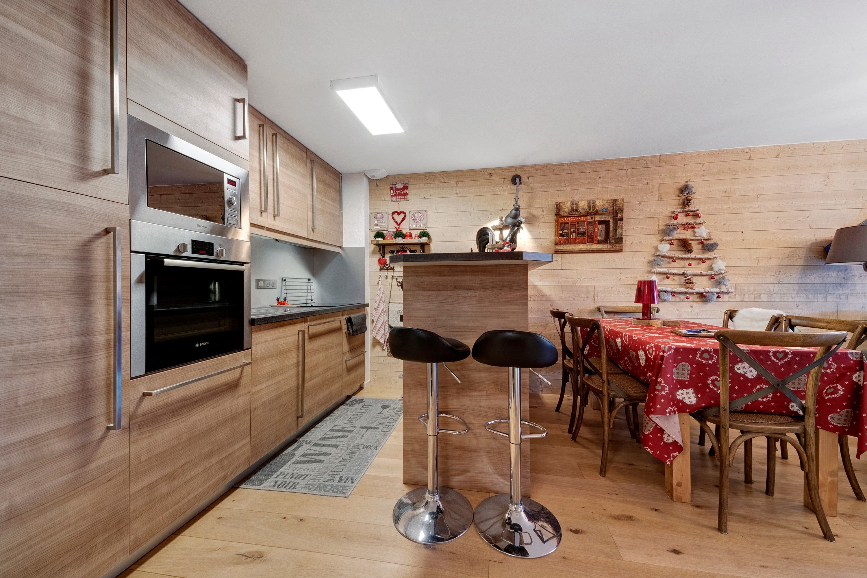 Une cuisine moderne avec des meubles en bois, un four Bosch et un coin repas agrémenté d’une nappe rouge.