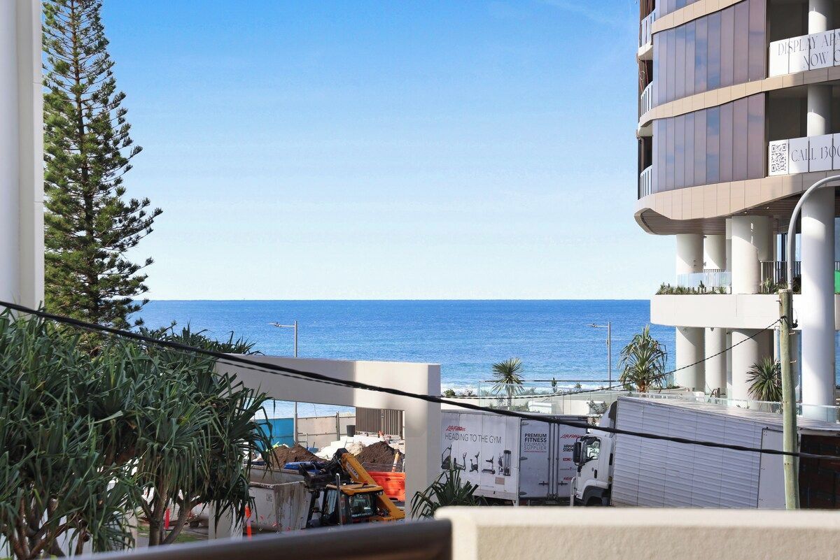 A view of a building with a balcony overlooking the sea, construction equipment, and a truck parked in front.