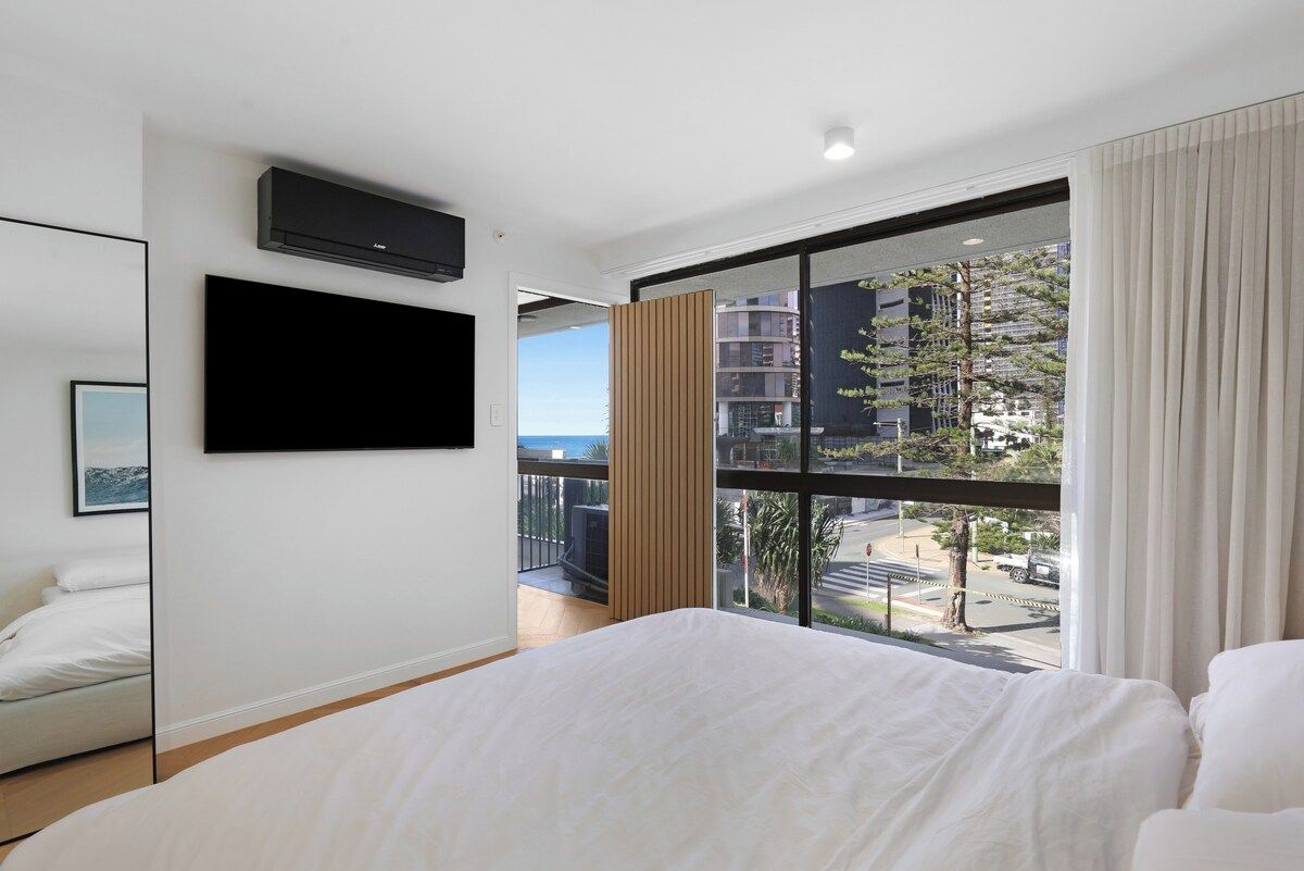 A hotel room with a large bed, a mounted TV, a window with a view of buildings and a street, and a balcony.