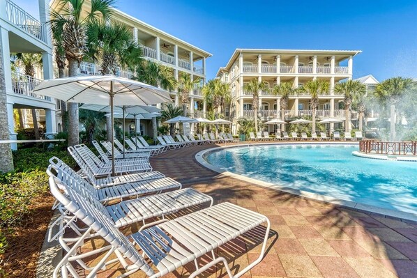 A resort pool area with sun loungers and umbrellas, surrounded by multi-story buildings with balconies. - Palms by the Sea Carriage House | Community Pool | Pets Welcome (Seacrest Beach)
