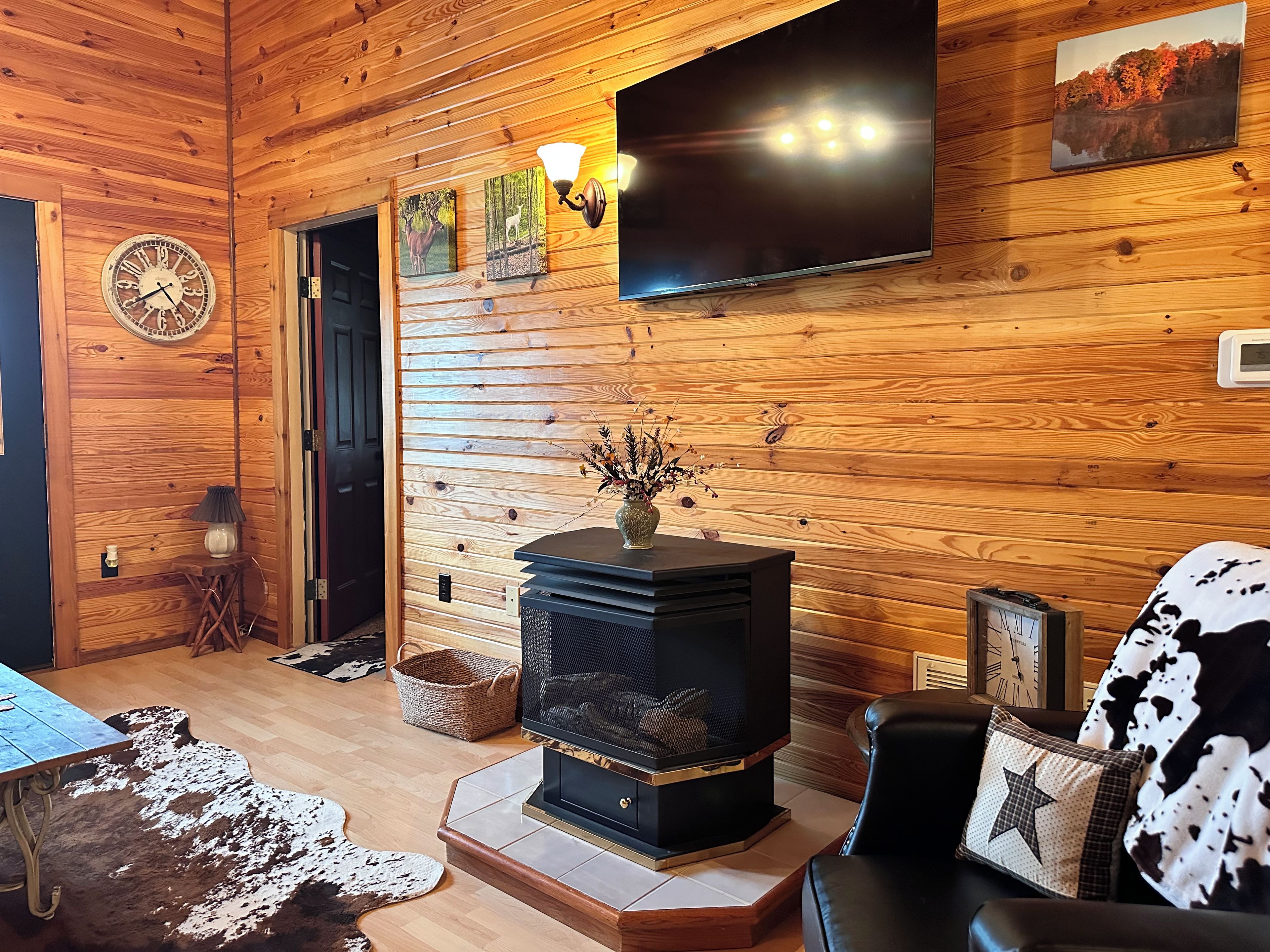 Un interior acogedor de cabaña con chimenea, paredes de madera, televisor montado y un reloj.