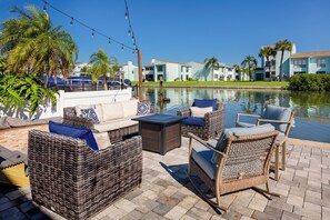 Un espace aménagé au bord de la piscine, avec des meubles en osier, une table et une vue sur un immeuble d’habitation situé de l’autre côté de l’eau.