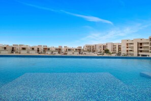 Ein Swimmingpool mit einer gefliesten Kante, umgeben von modernen Gebäuden mit Balkonen und einem strahlend blauen Himmel.