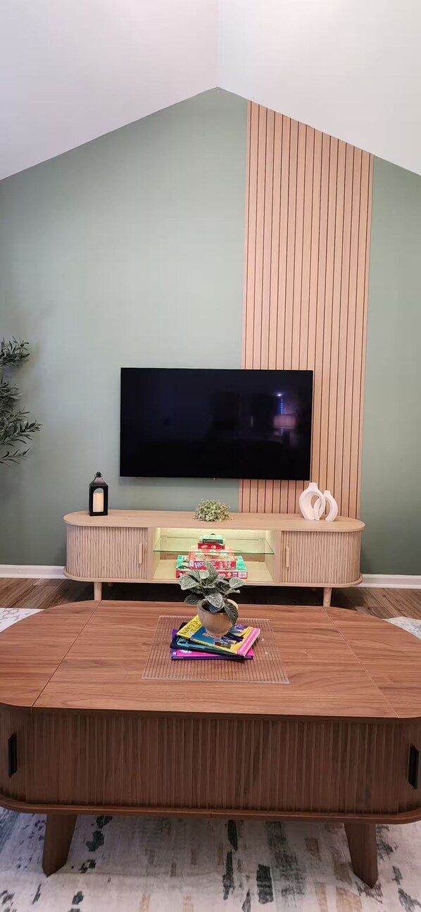 A modern living room with a large flat-screen TV mounted on a wooden wall, a round wooden coffee table with magazines and a plant, and a small side table with a candle and decorative items. - Modern 2BR - King Bed - Pet-Friendly - Sleeps 6 (Boiling Springs)