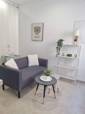 A modern living room with a grey sofa, a small round table with a plant, a white bookshelf, and a framed picture on the wall.