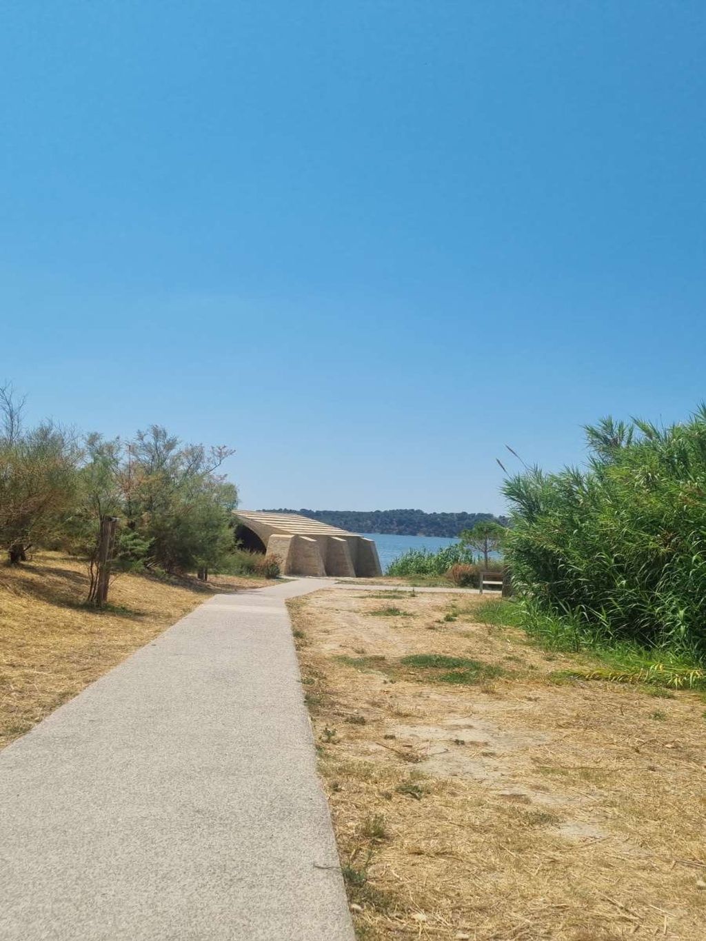 A paved pathway leading to a stone bridge over a body of water, surrounded by dry grass and sparse vegetation.
