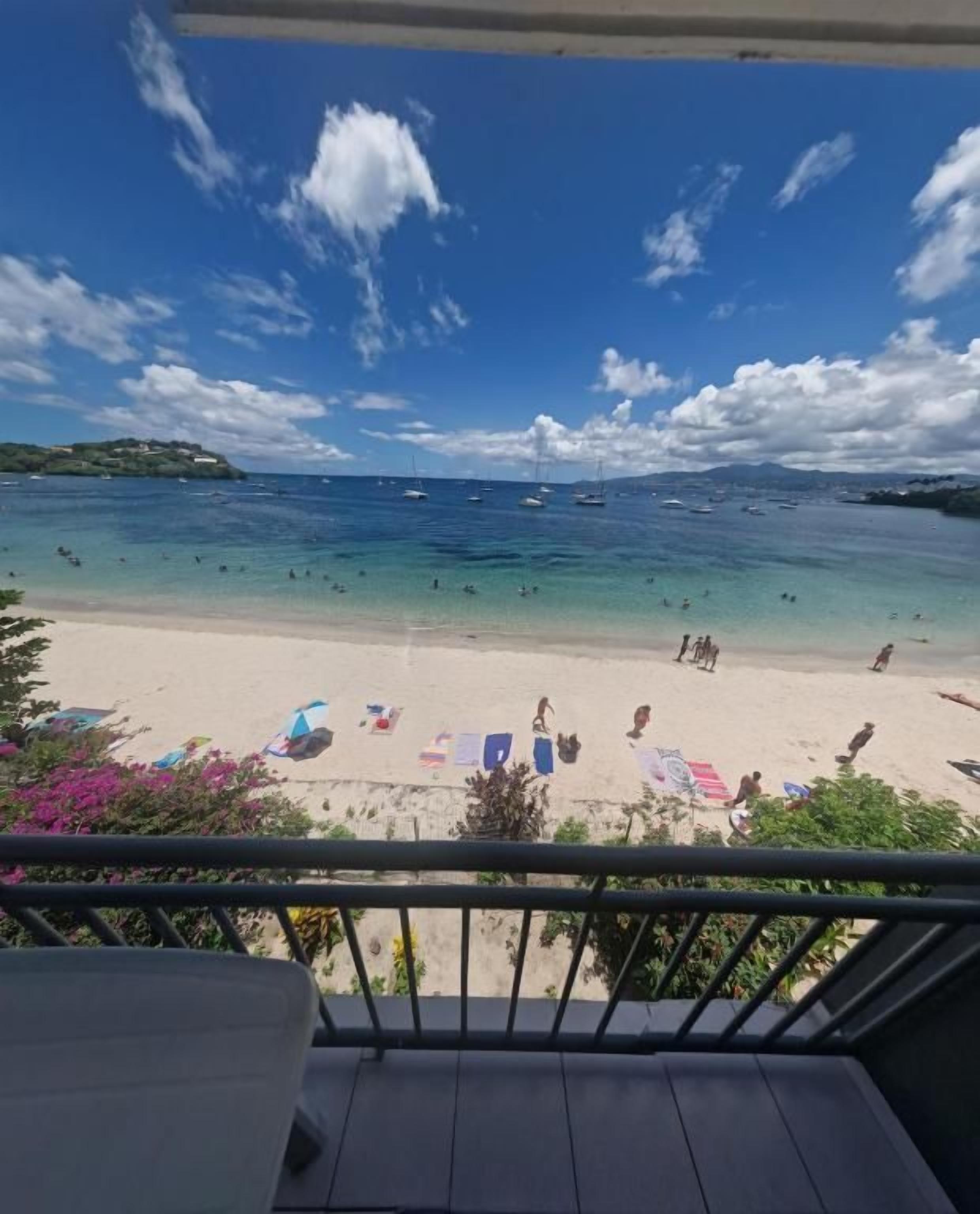 A beach view from a balcony with a railing, clear sky, and boats in the water.