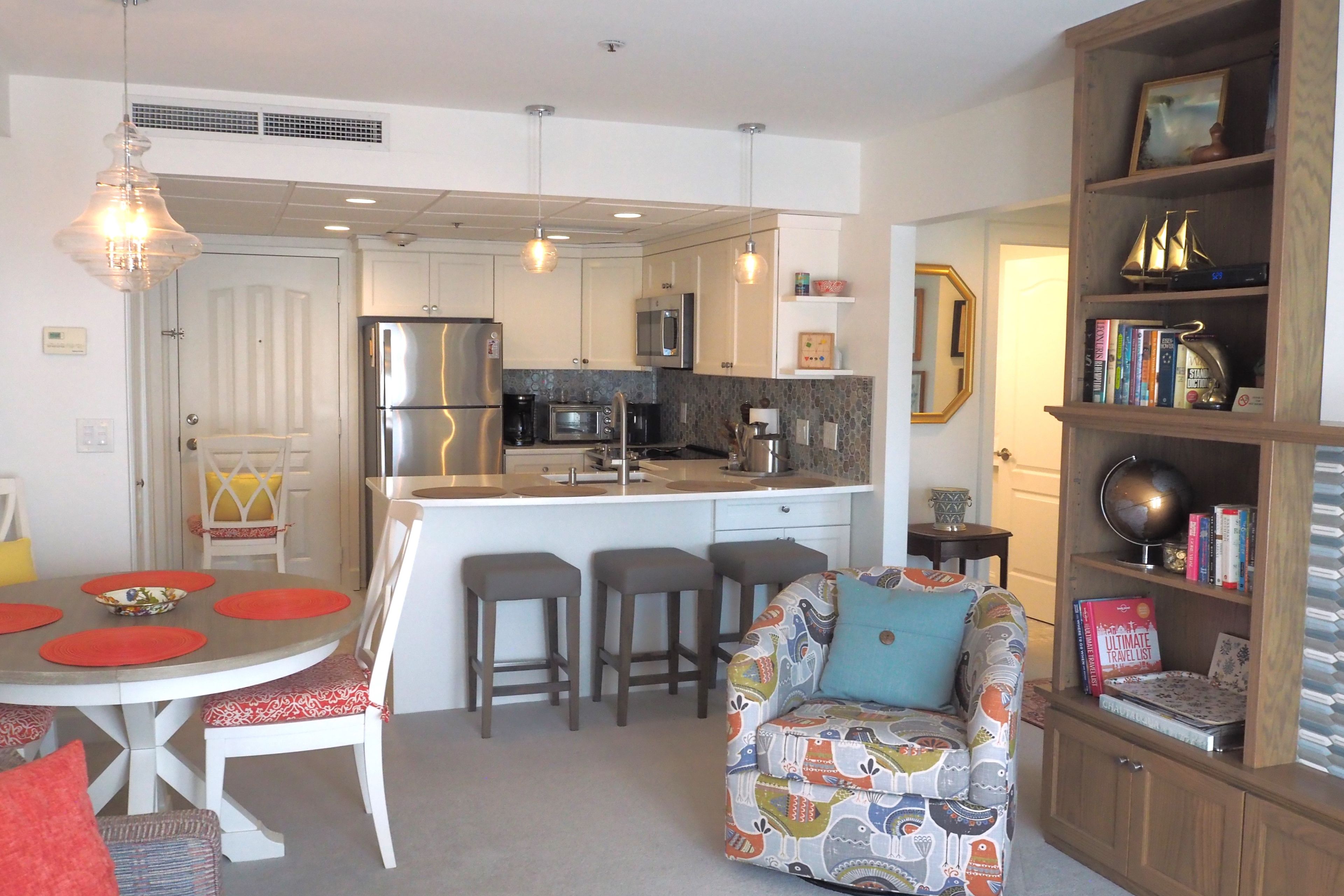 A modern kitchen with a central island, bar stools, a dining table with red placemats, and a bookshelf with books and decorative items.
