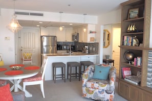 A modern kitchen with a central island, bar stools, a dining table with red placemats, and a bookshelf with books and decorative items.