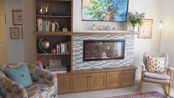 Una sala de estar con chimenea, televisor montado encima, estantería, sofá estampado y una silla decorativa.