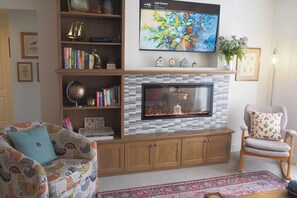 Ein Wohnzimmer mit Kamin, einem darüber montierten Fernseher, einem Bücherregal, einem gemusterten Sofa und einem dekorativen Sessel.