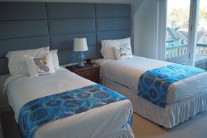 A hotel room with two beds, a nightstand, a lamp, and a window with a view of buildings.