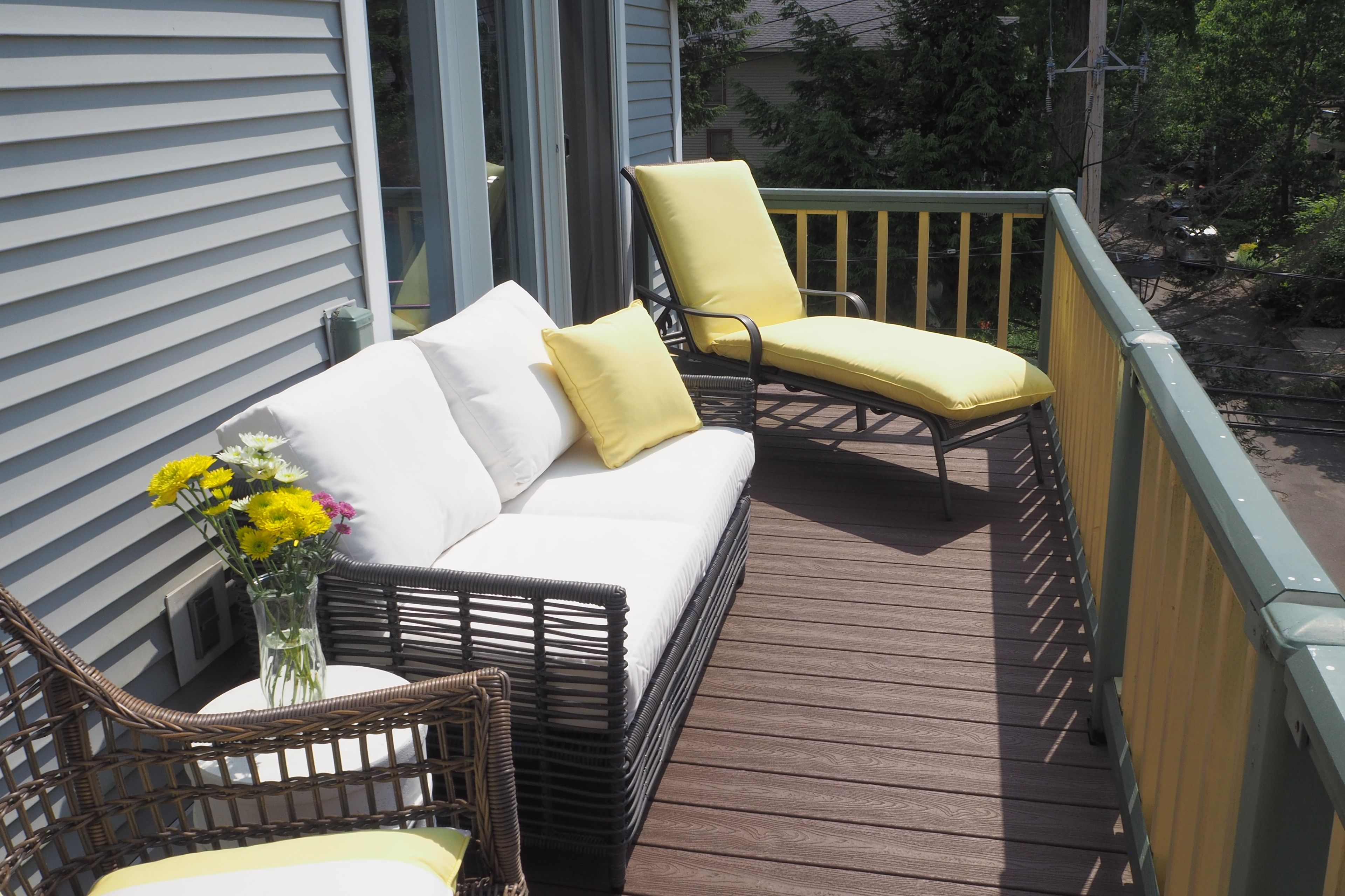 Una terraza de madera con un sofá blanco y dos sillas amarillas, una silla de mimbre y un jarrón con flores.