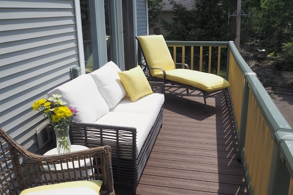 A wooden deck with a white sofa and two yellow chairs, a wicker chair, and a vase with flowers.