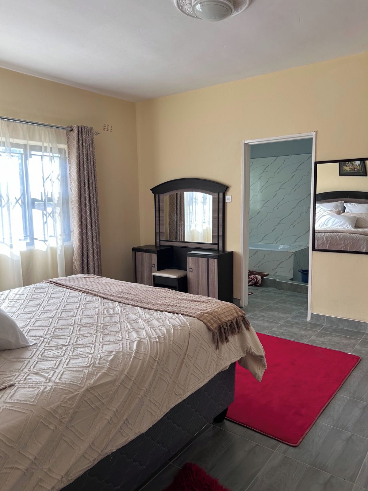 A bedroom with a bed, a desk, a mirror, and a bathroom visible through an open door.