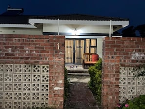 Una casa de ladrillo con entrada protegida por una reja, un porche con una silla y un pequeño sendero que lleva a la puerta.