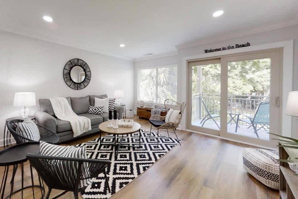 Un salon moderne avec un canapé, une table basse et des fauteuils, un tapis à motifs et une grande fenêtre donnant sur des meubles d’extérieur.