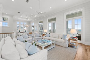 Un salon spacieux, doté de meubles blancs, de grandes fenêtres et d’un ventilateur de plafond.