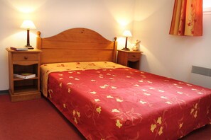 A neatly made bed with a floral bedspread, two bedside lamps, a nightstand with a book, and a wall-mounted heater. - T2 au pied remontées mécaniques | Piscine chauffee (Ax-les-Thermes)
