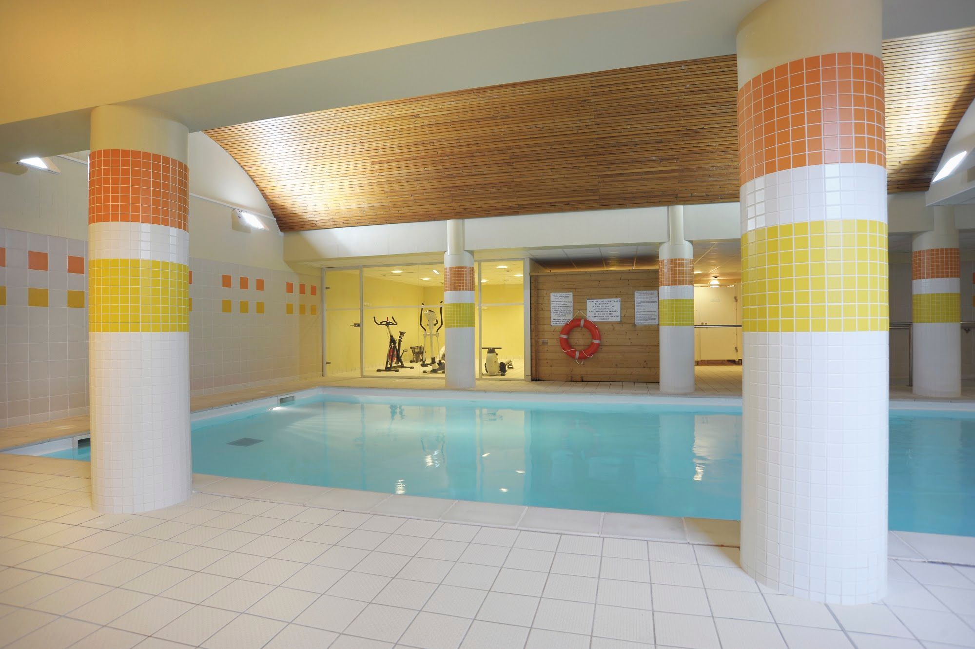 Une piscine intérieure aux murs et aux colonnes recouverts de carreaux, équipée de matériel d’exercice et dotée d’un plafond en bois.