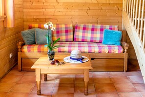 A wooden sofa with colorful cushions, a small table with a plant and sunglasses, and a wooden railing. - Balcon vue montagne | Piscine chauffée | Sauna (Morillon)