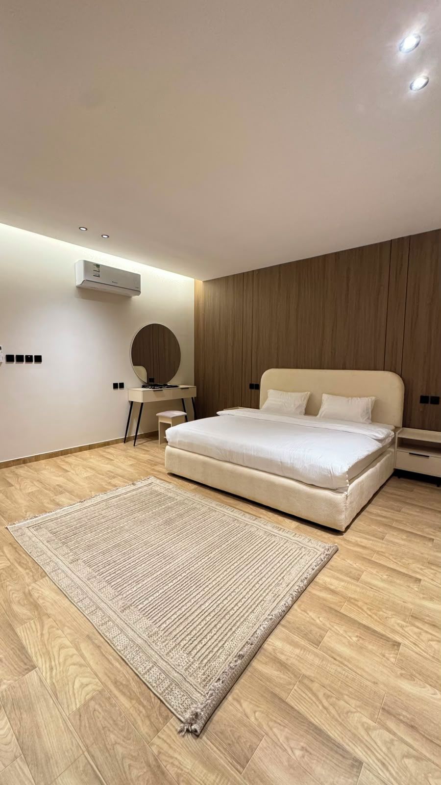 A modern bedroom with a large bed, a round mirror, a small table, and a wall-mounted air conditioner.