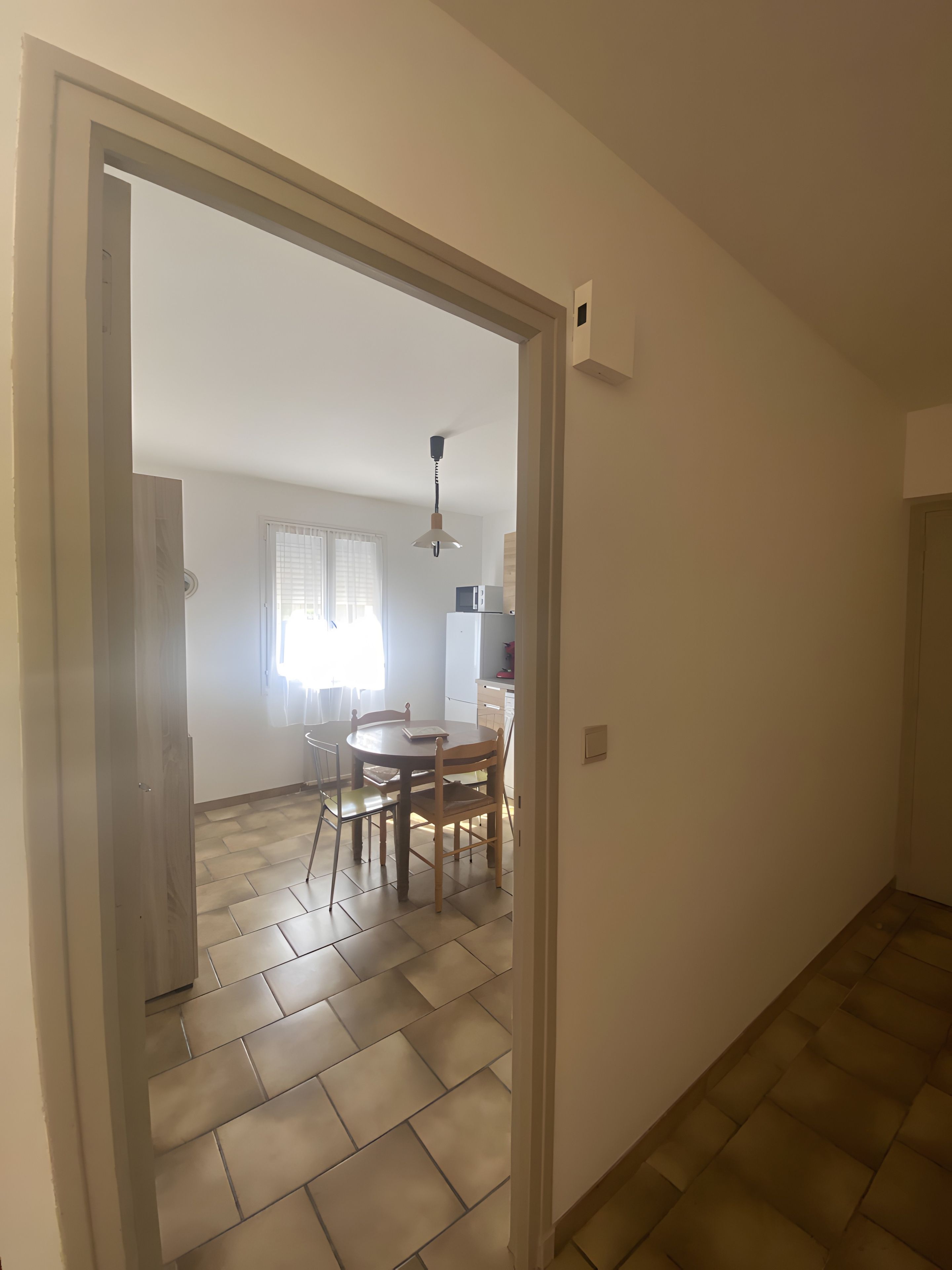 Un couloir mène à une salle à manger équipée d’une table et de chaises, d’un ventilateur de plafond et d’un réfrigérateur.