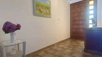 Une pièce avec un sol carrelé, un mur blanc orné d’un tableau encadré, une petite table sur laquelle est posé un vase de fleurs et une porte en bois.