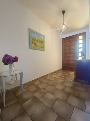A room with a tiled floor, a white wall with a framed painting, a small table with a vase of flowers, and a wooden door. - Cottage 'Gîte Tournesols' with Private Terrace, Wi-Fi and Air Conditioning (Arles-sur-Tech)