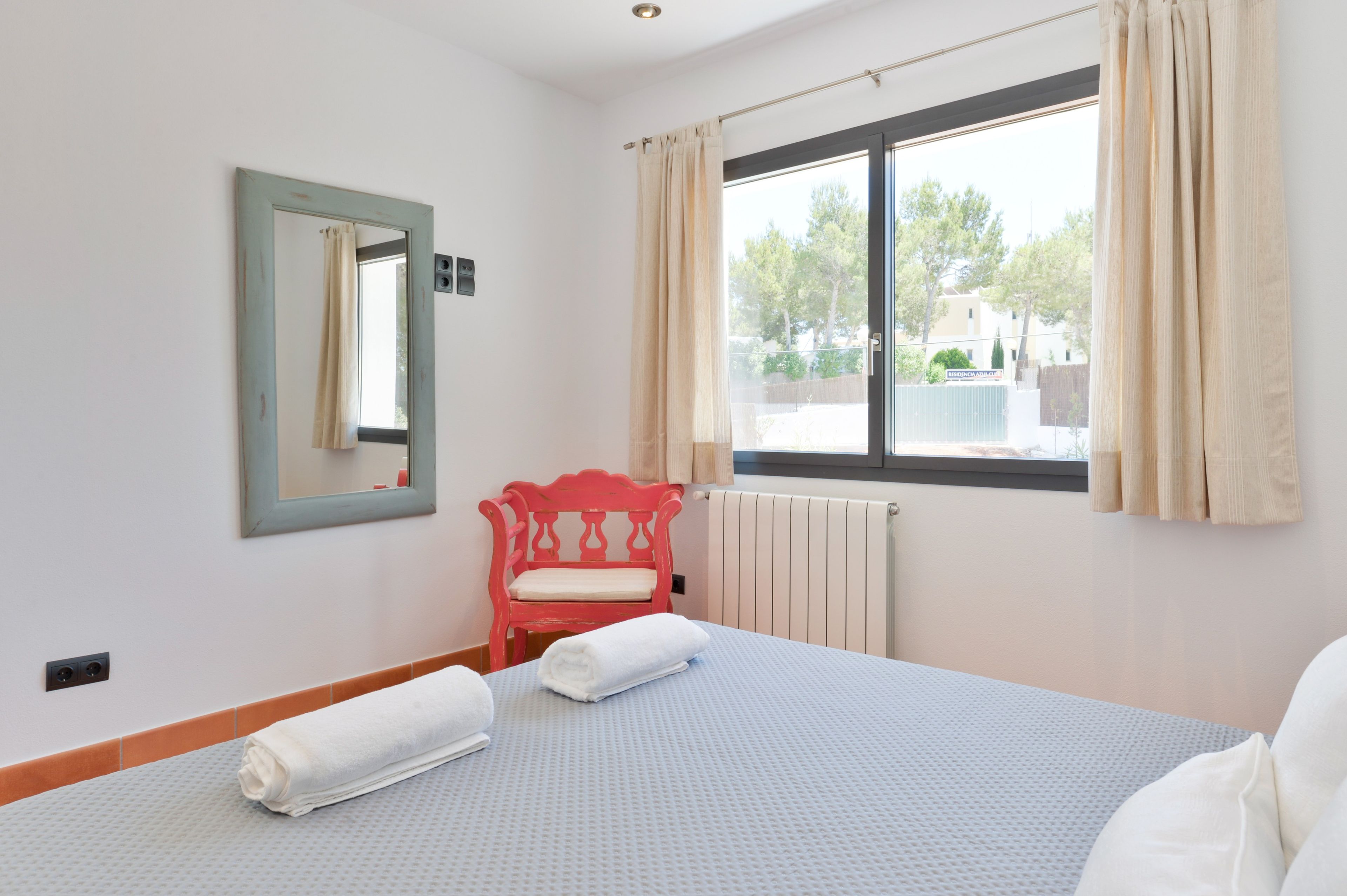 Una habitación de hotel con una cama, una silla roja, una ventana con cortinas, un radiador y un espejo en la pared.