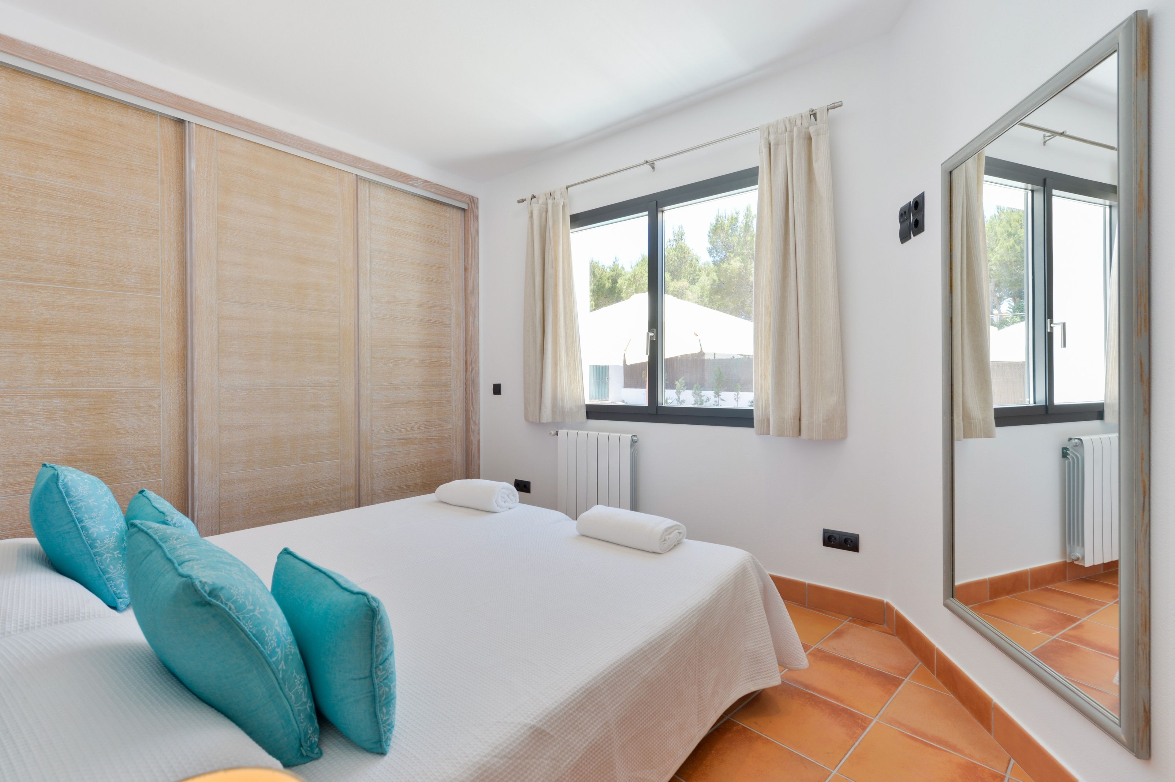 A hotel room with a bed, two pillows, a window with curtains, a radiator, and a large mirror.