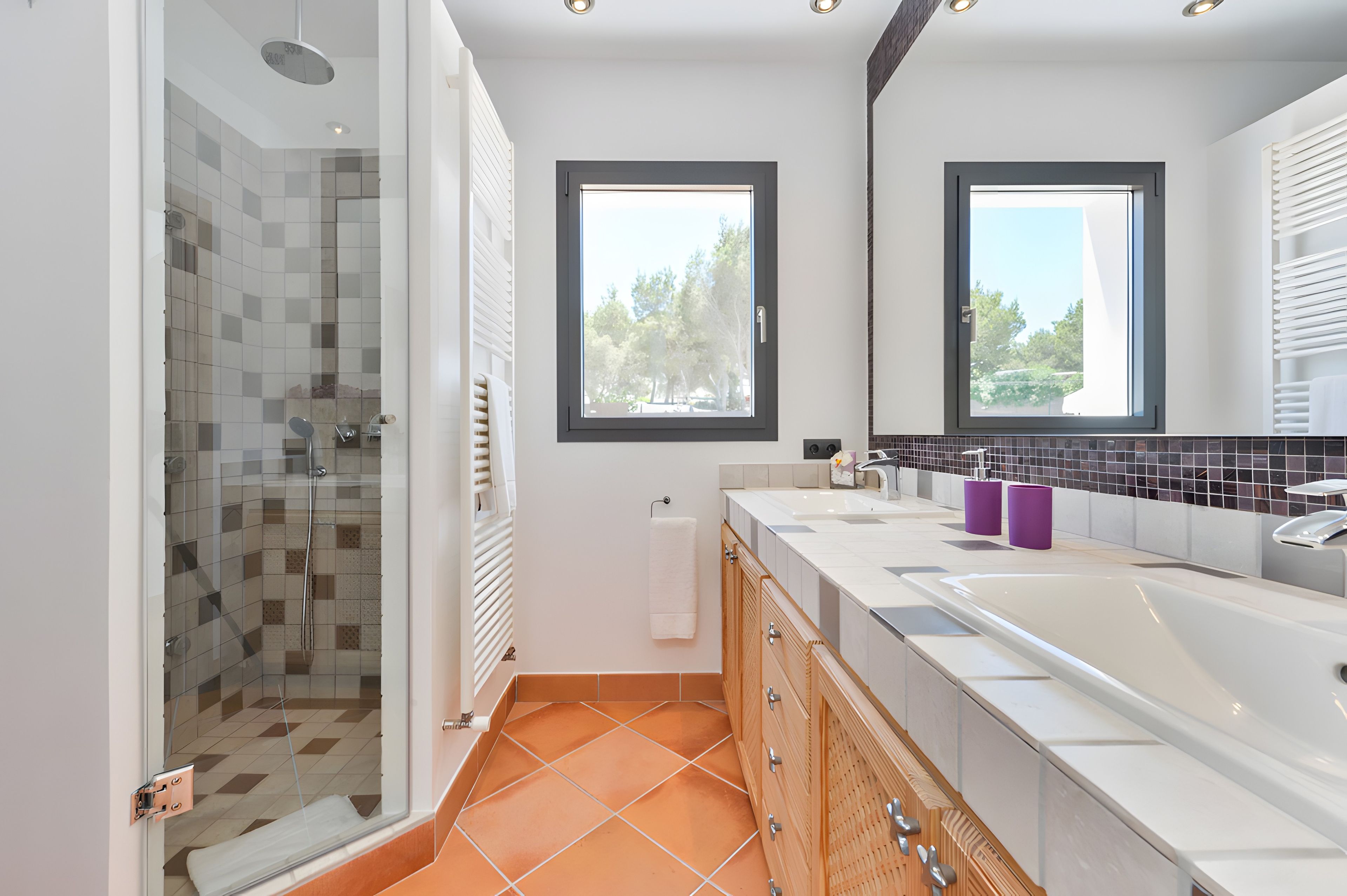 Une salle de bain moderne dotée d’une douche à parois de verre, d’une grande baignoire et de deux fenêtres qui laissent entrer la lumière naturelle.