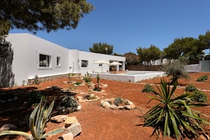 Un bâtiment moderne de couleur blanche, doté d’une piscine, et entouré d’un jardin aménagé avec différentes plantes et rochers.
