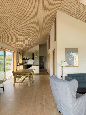 A spacious living area with a wooden ceiling, a dining table with chairs, a sofa, and a kitchen in the background. - Architect-designed house on Møn close to beach (Stege)