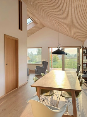Ein geräumiger Essbereich mit langem Tisch und Stühlen, eine hohe Decke mit Holzbalken und große Fenster mit Blick ins Freie.