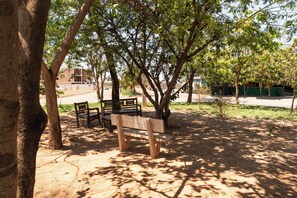 A park with a bench under a tree, a paved path, and a building in the background. - Hotel do Cerrado (Luís Eduardo Magalhães)
