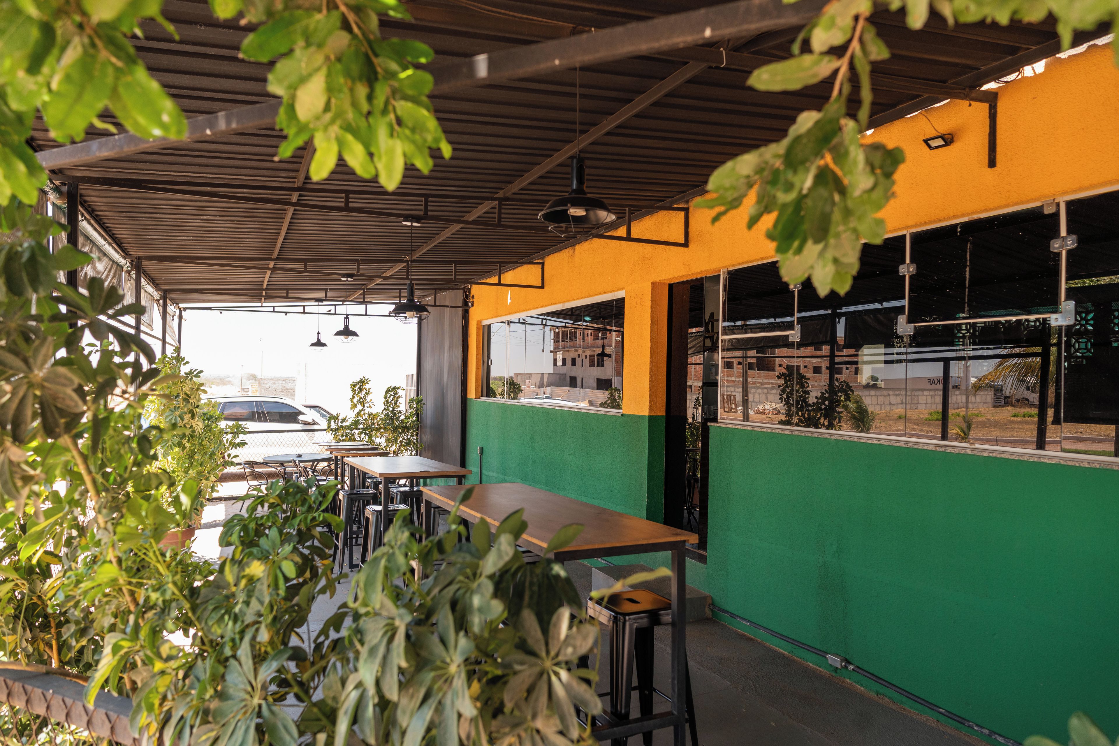 An outdoor seating area with tables and chairs, surrounded by greenery and a metal roof structure.