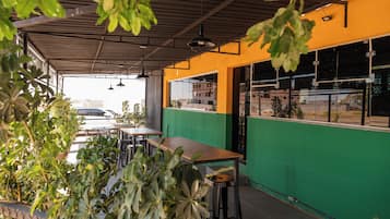An outdoor seating area with tables and chairs, surrounded by greenery and a metal roof structure.
