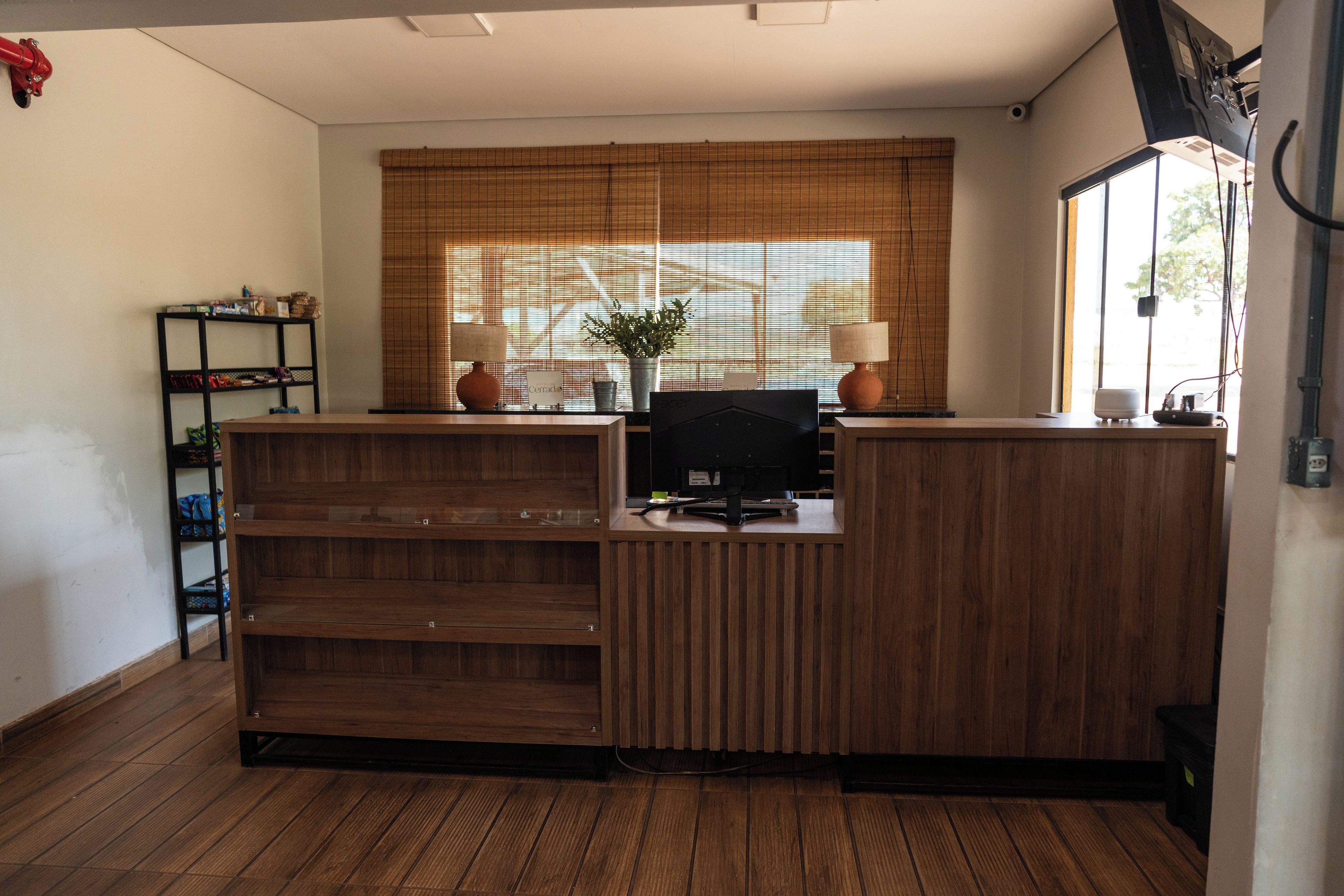 A reception desk with a computer, a potted plant, and a window with blinds.