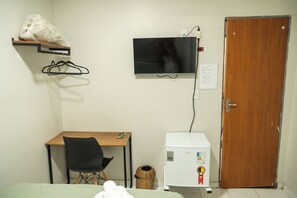 Une petite pièce comprenant un bureau, une chaise, un téléviseur fixé au mur, un réfrigérateur et une porte en bois.