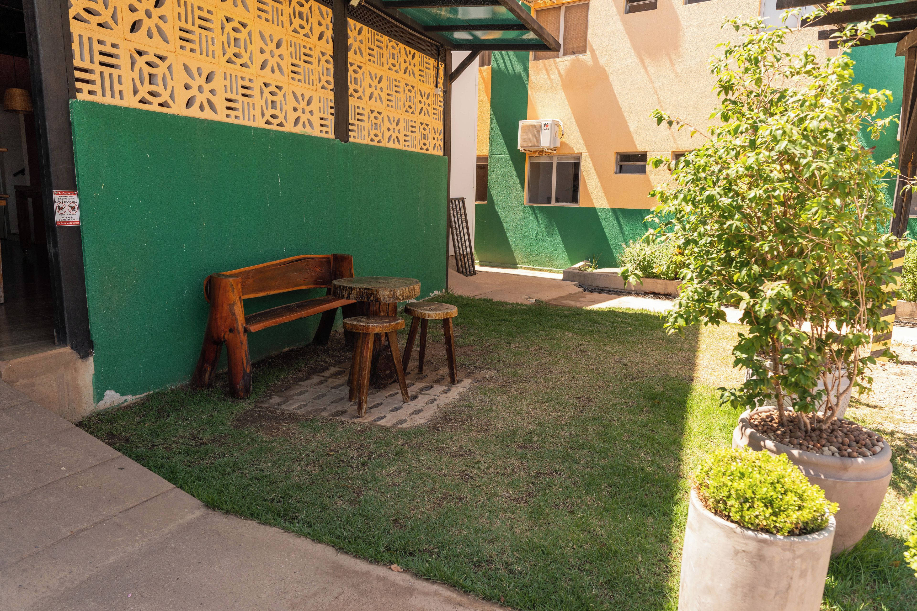 A small outdoor seating area with a bench and two stools, a potted plant, and a green wall with a decorative pattern.