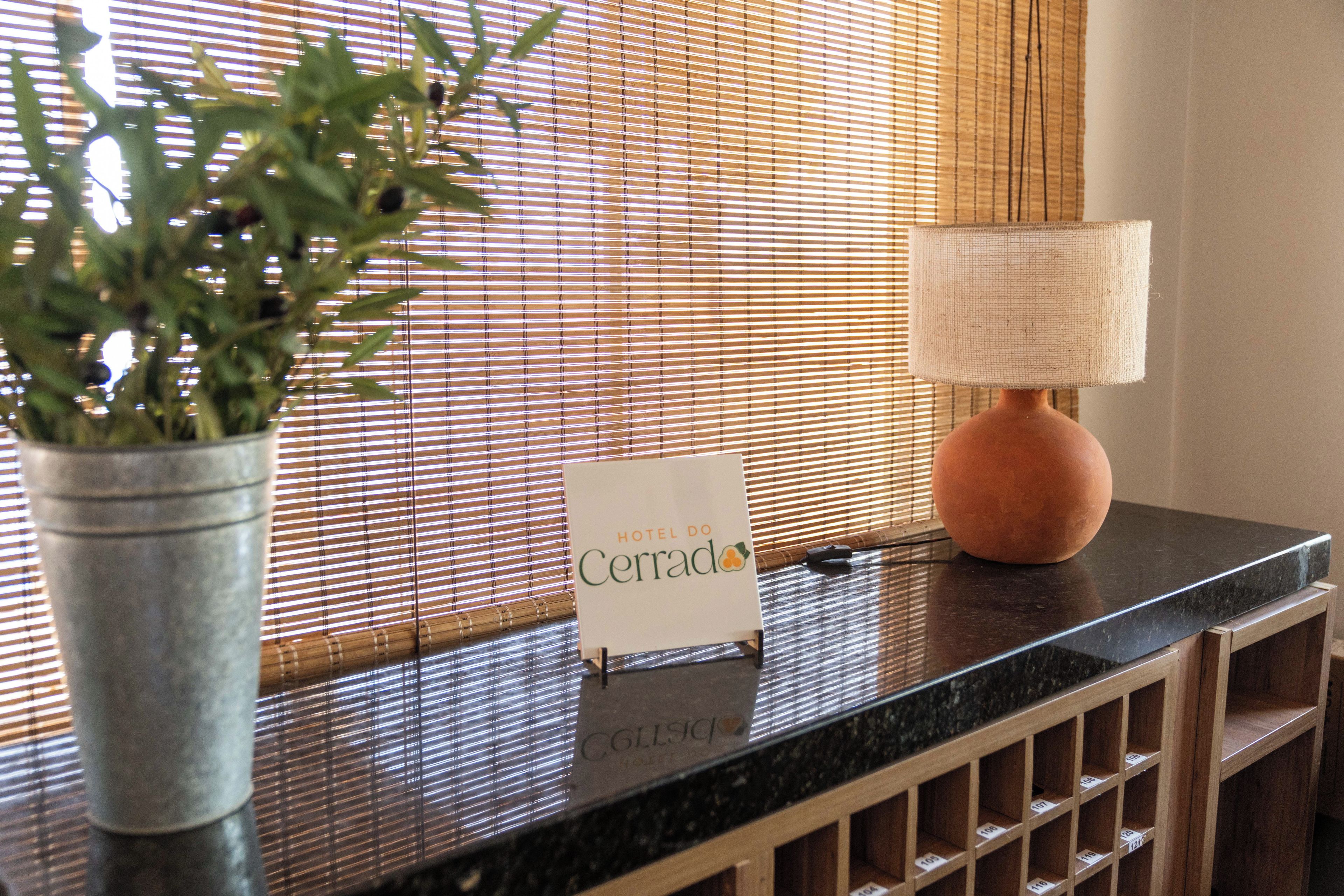 A reception desk with a sign, a potted plant, a lamp, and a window with blinds.